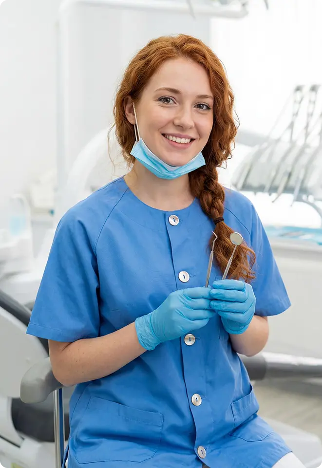 A smiling female dentist in blue scrubs holding dental instruments in a modern dental clinic