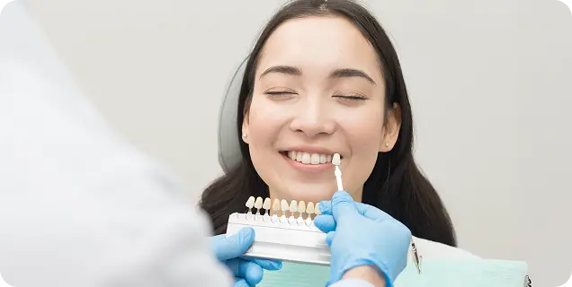 A dentist matching tooth shade samples to a smiling female patient's teeth at a dental clinic