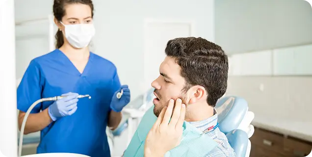 A male patient holding his jaw in pain while a dentist in blue scrubs prepares dental instruments nearby