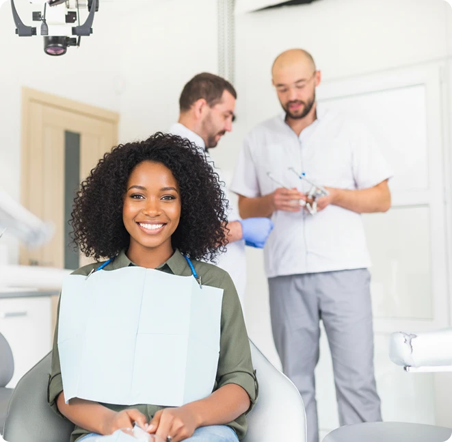 Smiling female patient in a dental bib sitting in a dental chair with two dentists preparing instruments in the background