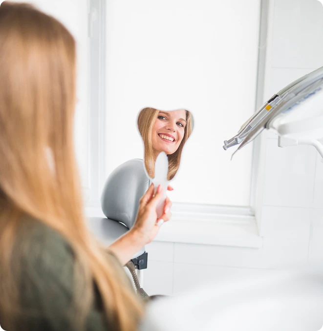 Female patient smiling at her reflection in a tooth-shaped mirror while sitting in a dental chair