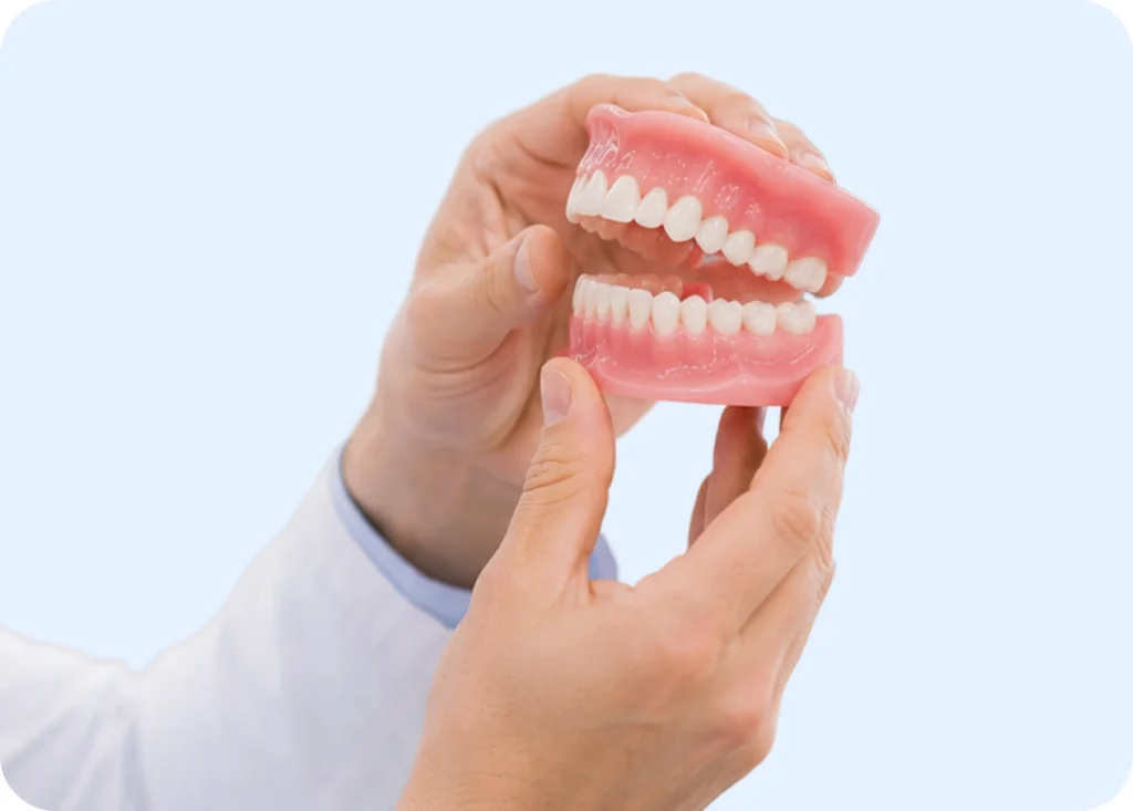 Hands holding a complete set of full upper and lower dentures against a light blue background